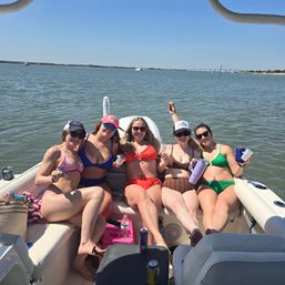 Five friends in colorful bikinis smiling and holding drinks while relaxing on a pontoon boat in a sunny coastal bay with calm water and a distant bridge