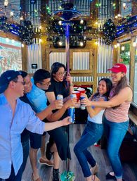 Six friends laughing and toasting with beer cans and red cups inside a festively lit party trolley with a center dance pole, string lights and wooden interior.
