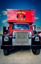 Front-facing red retro food truck with shiny chrome grille, round headlights, and a colorful rooftop sign featuring a cartoon rooster and musical notes, parked outdoors under a bright blue sky.