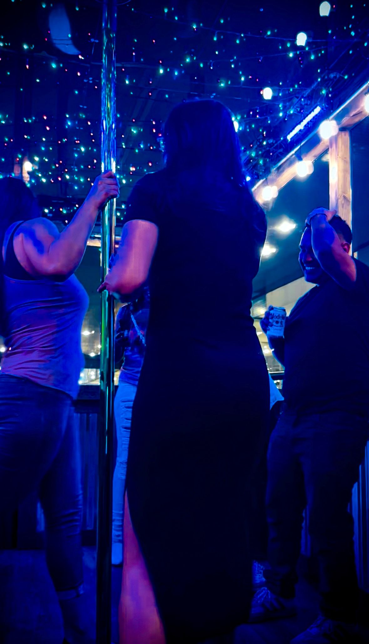 Friends dancing around a chrome pole under blue neon and twinkling string lights on a rooftop patio at night, one person holding a canned drink.