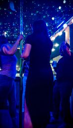 Friends dancing around a chrome pole under blue neon and twinkling string lights on a rooftop patio at night, one person holding a canned drink.