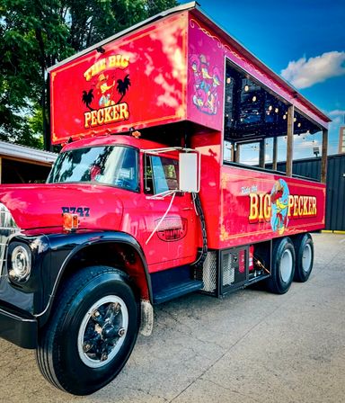 Bright red vintage truck converted into a mobile party bar with open-sided upper deck, colorful cartoon rooster mural, string lights, parked outdoors under a blue sky