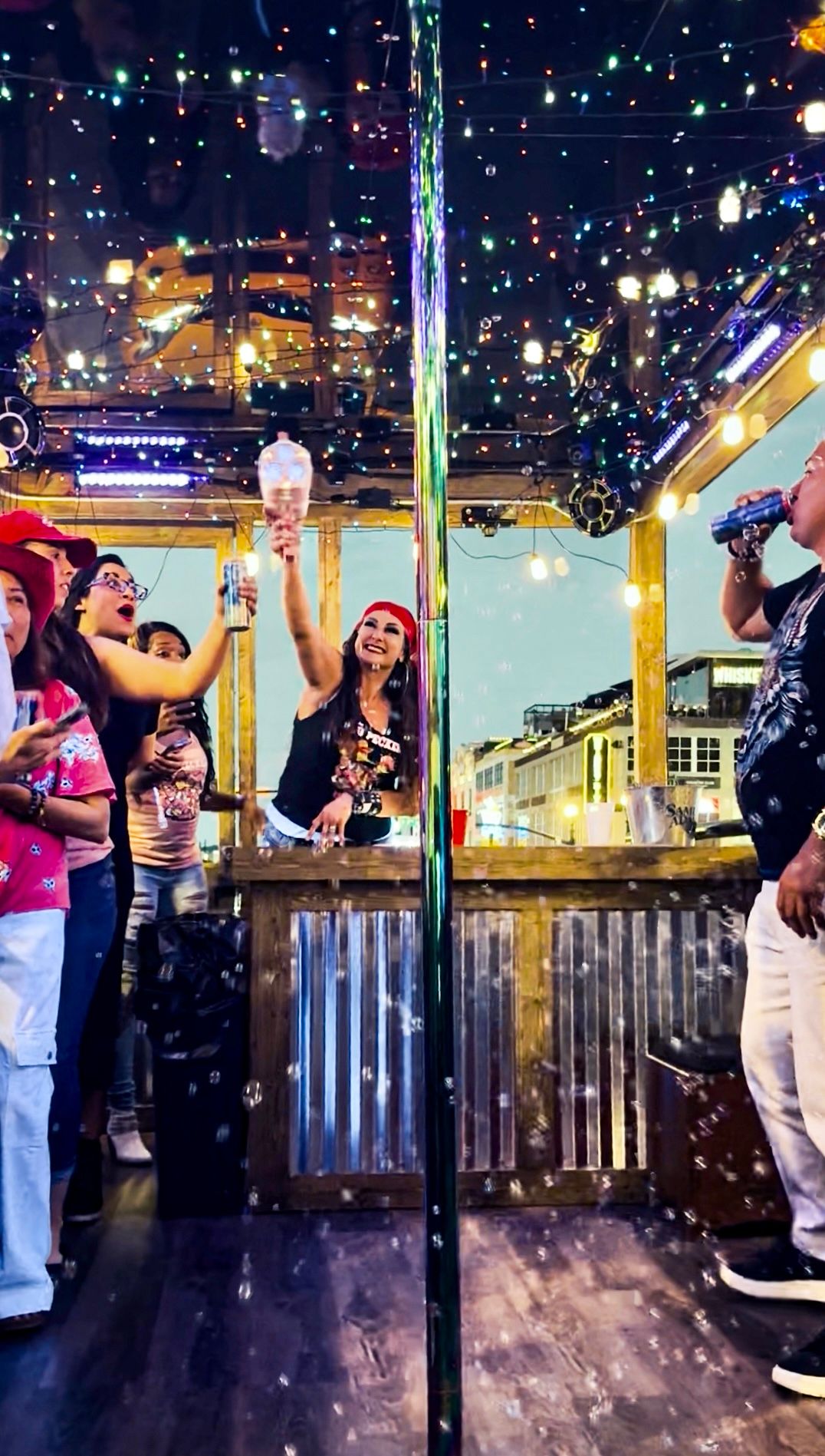 Partygoers cheering at a lively downtown bar patio at night — woman in a red bandana raises a bottle behind a wooden, corrugated-metal bar, colorful string lights reflected on a mirrored ceiling, bubbles floating and city buildings visible through the windows.