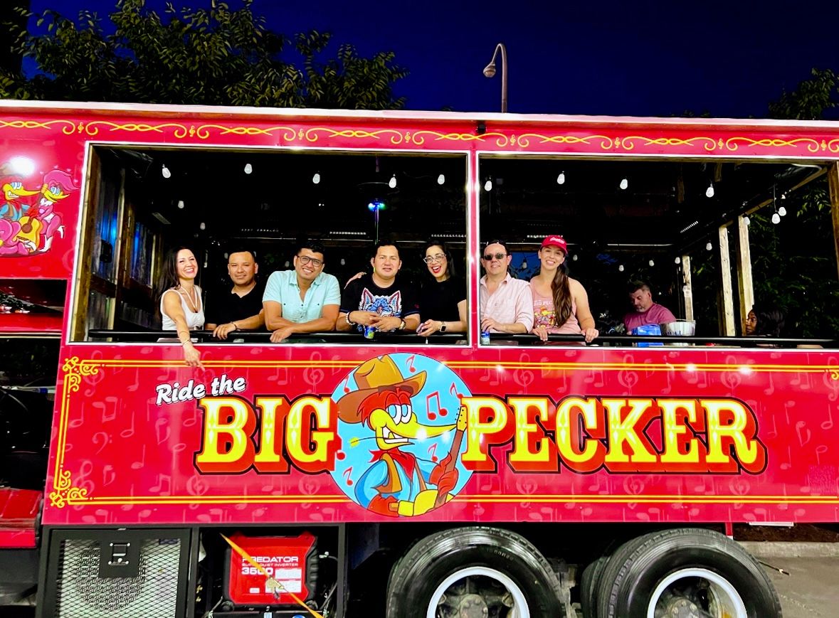 Smiling group of adults leaning from the windows of a bright red party trolley at night, decorated with string lights and a colorful cartoon woodpecker logo and bold yellow lettering.