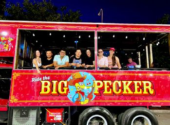 Smiling group of adults leaning from the windows of a bright red party trolley at night, decorated with string lights and a colorful cartoon woodpecker logo and bold yellow lettering.