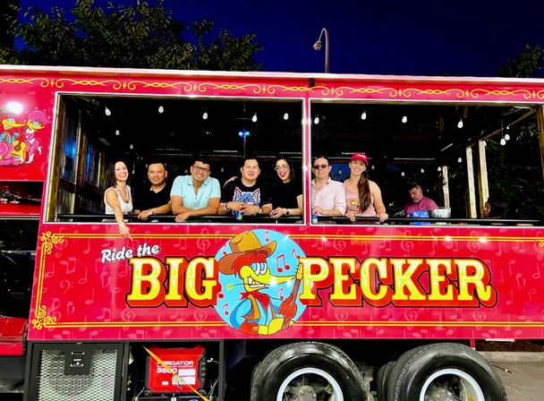 Smiling group of adults leaning from the windows of a bright red party trolley at night, decorated with string lights and a colorful cartoon woodpecker logo and bold yellow lettering.