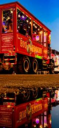 Vibrant red open-sided party truck with colorful string lights and passengers on a lively downtown street at dusk, its illuminated side reflected in a puddle.
