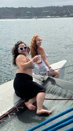 Two friends in sunglasses and swimsuits sitting on a catamaran bow net, smiling and holding drinks with calm ocean water and a distant shoreline in the background.