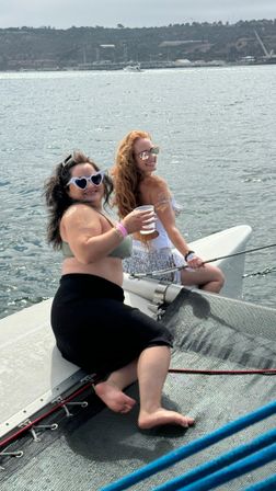 Two friends in sunglasses and swimsuits sitting on a catamaran bow net, smiling and holding drinks with calm ocean water and a distant shoreline in the background.