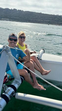 Two smiling adults in sunglasses sitting on a sailboat with legs dangling over sparkling coastal bay water and a hilly shoreline in the background