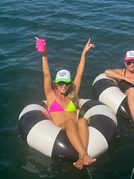 Woman in neon pink-and-green bikini and 'Forty AF' trucker hat lounging on black-and-white inflatable tube in open water, holding a pink cup and raising her arms in celebration.