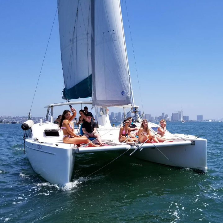 White catamaran sailing on bright blue bay water with a group of people in swimsuits lounging on the front trampoline, coastal city skyline in the distance under a clear sky.
