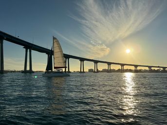 Sailboat gliding beneath a long concrete bridge at golden sunset, sun sparkling on bay waters with wispy clouds