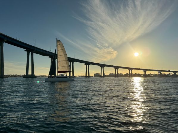 Sailboat gliding beneath a long concrete bridge at golden sunset, sun sparkling on bay waters with wispy clouds