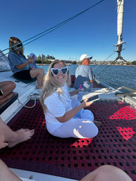 Smiling person in heart-shaped sunglasses sitting on a sailboat net, holding a phone and drink with a sunny coastal harbor and shoreline in the background.
