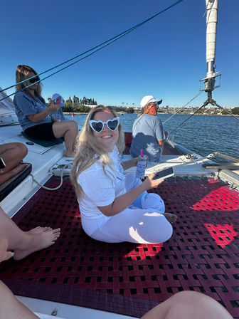 Smiling person in heart-shaped sunglasses sitting on a sailboat net, holding a phone and drink with a sunny coastal harbor and shoreline in the background.