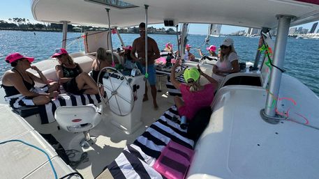 Group enjoying a sunny harbor sail on a catamaran-style boat, lounging on navy-and-white striped towels, several wearing bright pink hats and a neon cap, with a coastal city skyline and palm-lined shore in the background.