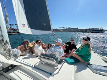 Group of friends lounging on a sailboat under full sail on a sunny day, sparkling blue water and a palm-lined coastal harbor with a pier in the background.