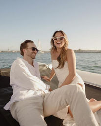 Happy couple in white outfits and sunglasses lounging on a yacht on a sunny bay with sailboats and a distant shoreline