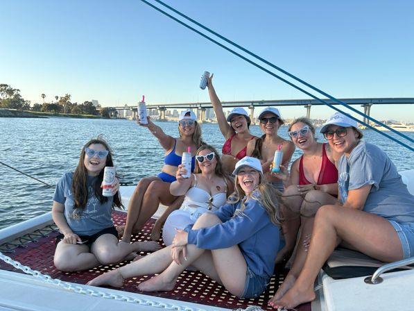 Eight friends laughing and raising canned drinks while lounging on a sailboat trampoline, with a bridge and city skyline over a calm bay on a sunny evening.