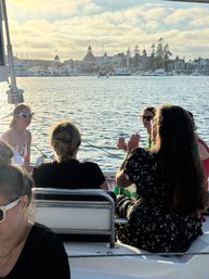 Women enjoying a leisure boat cruise in a sunny coastal harbor at sunset, chatting and sipping drinks with yachts and a historic waterfront hotel silhouette across the marina.