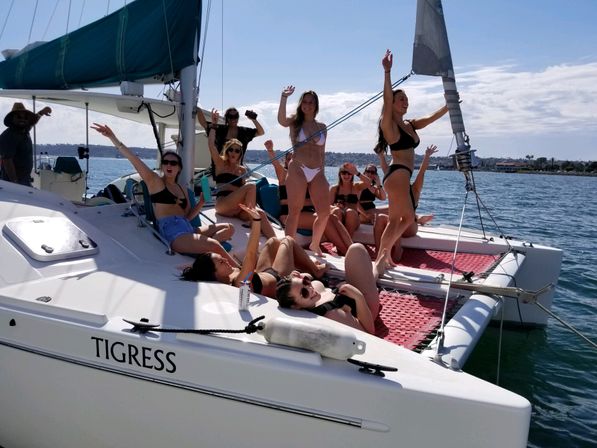 Group of women in swimsuits cheering and lounging on the foredeck and trampoline of a white catamaran on sunny coastal waters.
