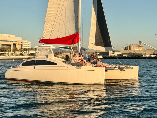 Sunlit catamaran cruising in a coastal marina at golden hour, passengers relaxing on deck with a red sail cover and waterfront buildings in the background.