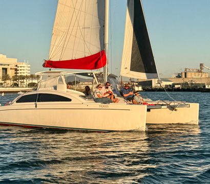 Sunlit catamaran cruising in a coastal marina at golden hour, passengers relaxing on deck with a red sail cover and waterfront buildings in the background.