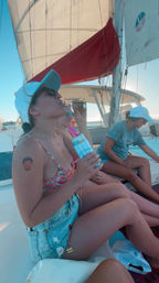 Three people relaxing on a sailboat in coastal waters at sunset — person in a floral bikini and denim shorts sipping from a bottle while holding a striped can cooler, others lounging in caps and sunglasses under white sails, summery sailing vibe.