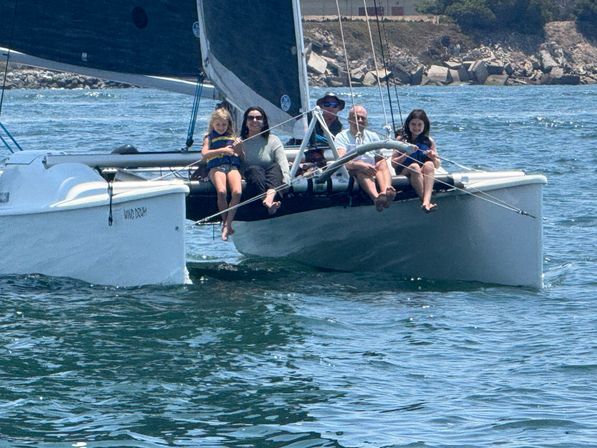 Group of five people lounging on the trampoline of a white catamaran, sailing on blue ocean water near a rocky coastline on a sunny day.