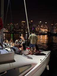 Friends relaxing on a sailboat at night with a glowing city skyline and a festoon‑lit tall ship reflecting across the harbor waters.