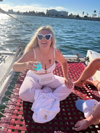 Smiling person in white 'BRIDE' fanny pack and heart-shaped sunglasses holding a bright blue cocktail while sitting on a catamaran net with a coastal city skyline and palm trees across the harbor