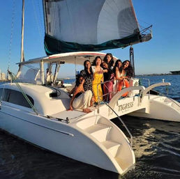 Smiling group of women on the stern of a white catamaran sailboat in colorful summer dresses, large sail overhead, cruising a calm coastal bay at golden hour