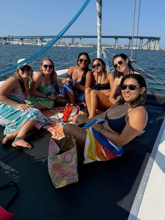 Group of friends in swimsuits relaxing on a sailboat trampoline, snacking and smiling with San Diego Bay and the Coronado Bridge visible in the background.