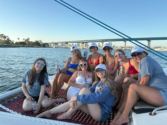 Smiling group of friends lounging on a sailboat trampoline with canned drinks, coastal city skyline and bridge in the background under a clear blue sky.