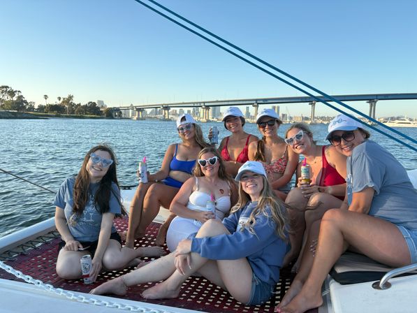 Smiling group of friends lounging on a sailboat trampoline with canned drinks, coastal city skyline and bridge in the background under a clear blue sky.