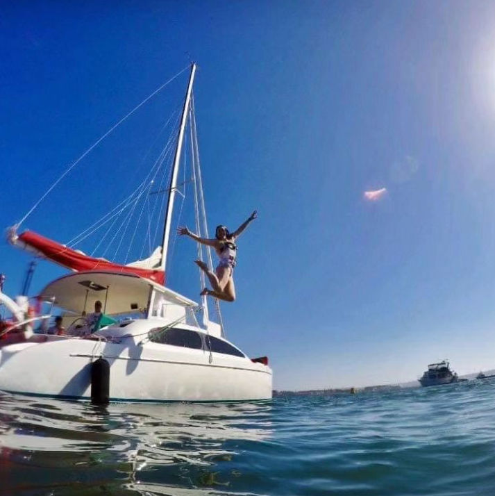 Person mid-air jumping off a white catamaran with a red sail cover into clear blue coastal water under a bright sunny sky — fun summer sailing scene.