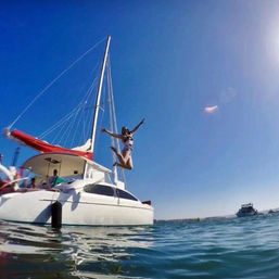 Person mid-air jumping off a white catamaran with a red sail cover into clear blue coastal water under a bright sunny sky — fun summer sailing scene.