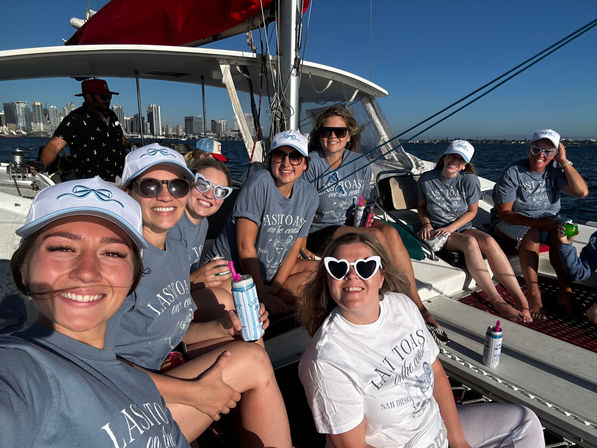Happy group of women in matching caps and t-shirts aboard a sailboat, holding drinks with the San Diego skyline and blue ocean on a sunny day