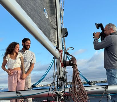 Smiling couple holding hands on a sailboat deck by the mast and coiled ropes while a photographer captures their portrait against a clear blue sky and distant coastal skyline.