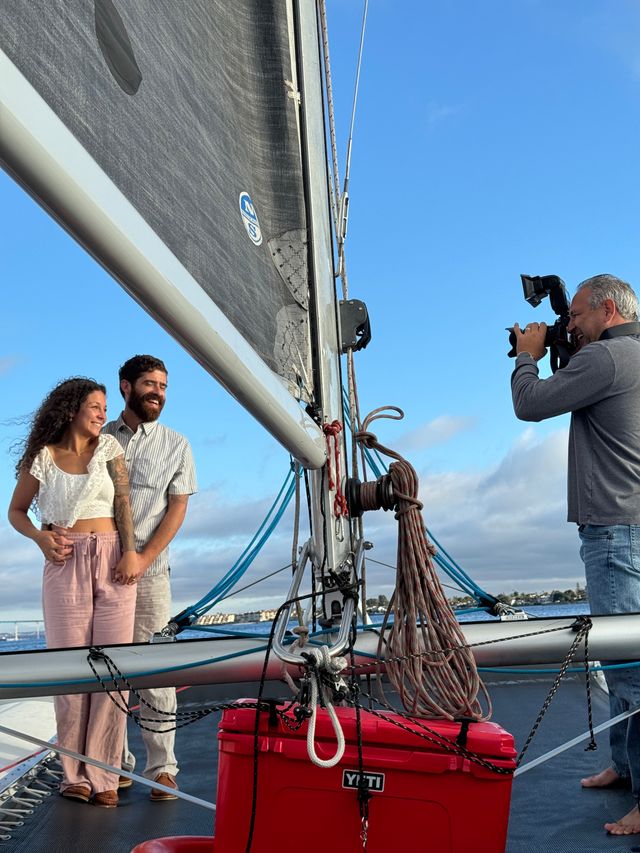 Smiling couple holding hands on a sailboat deck by the mast and coiled ropes while a photographer captures their portrait against a clear blue sky and distant coastal skyline.