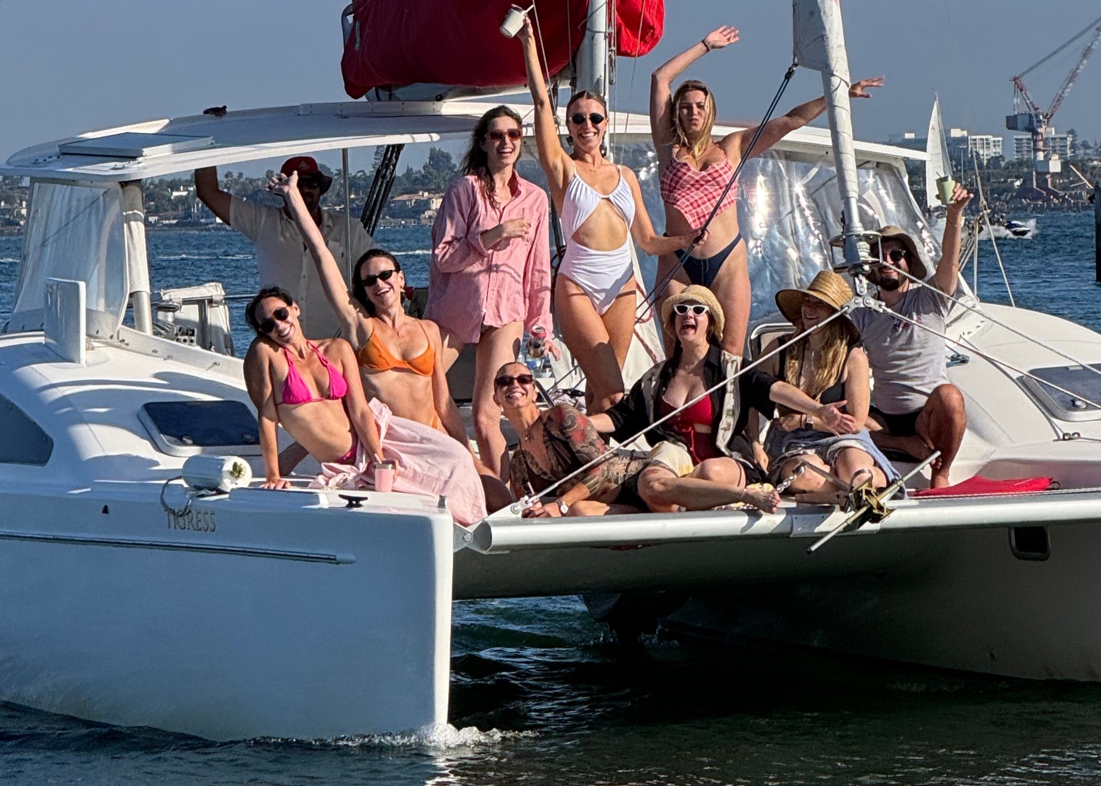 Group of friends in colorful swimsuits laughing and posing on the bow of a white catamaran sailboat, waving and holding drinks on a sunny summer day on coastal waters.