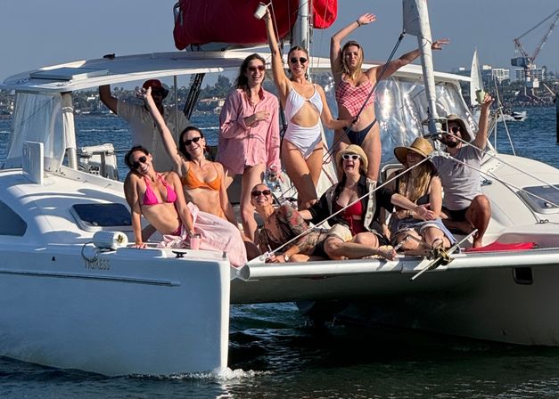Group of friends in colorful swimsuits laughing and posing on the bow of a white catamaran sailboat, waving and holding drinks on a sunny summer day on coastal waters.