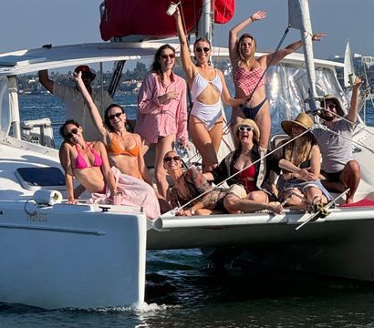 Group of friends in colorful swimsuits laughing and posing on the bow of a white catamaran sailboat, waving and holding drinks on a sunny summer day on coastal waters.