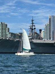 White catamaran sailboat with raised sails cruising past a massive docked naval aircraft carrier and city skyline under a sunny blue sky.