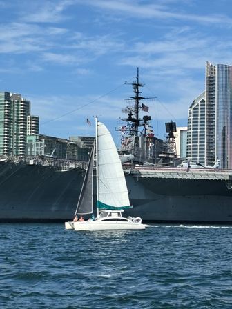 White catamaran sailboat with raised sails cruising past a massive docked naval aircraft carrier and city skyline under a sunny blue sky.