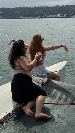 Two friends relaxing on the bow trampoline of a catamaran, one holding a cup with arms outstretched, overlooking a calm coastal bay and distant hills with a passing boat.