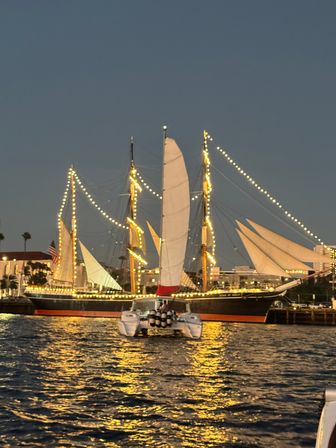 Dusk harbor scene: festively lit tall ship docked at a coastal marina with a white catamaran in the foreground and golden light reflections on the water.