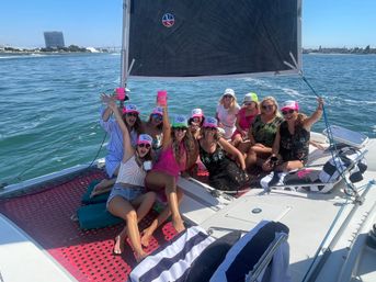 Women in colorful caps and sunglasses enjoying a sunny sailboat party in a calm bay, raising cups and smiling with a coastal city skyline in the background.