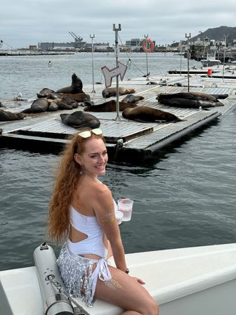 Smiling woman in a white swimsuit sits on a boat holding a drink while dozens of sea lions laze on floating docks at a foggy coastal harbor marina.
